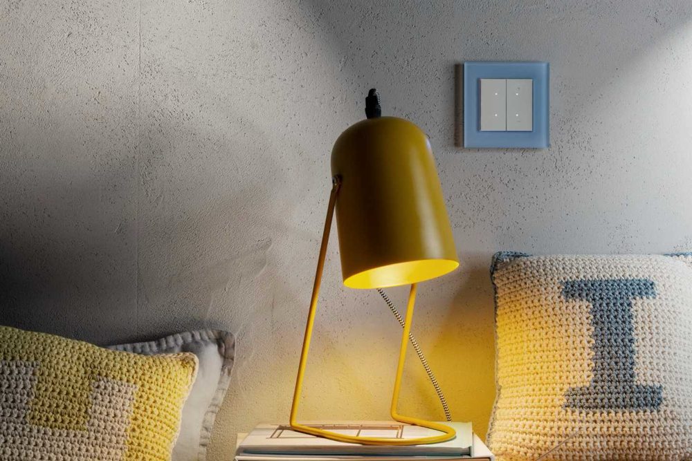 Biały sterownik z błękitną ramką SIMON GO na ścianie sypialni z żółta lampą – wygoda i bezpieczeństwo w polskim systemie inteligentnego domu. 