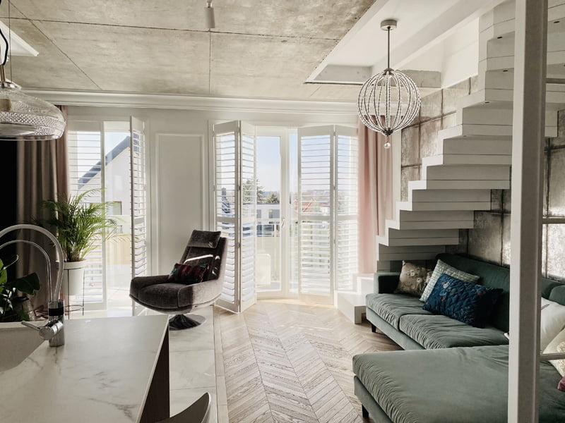 Nowoczesny salon z białymi okiennicami shutters wewnętrznymi przy dużych oknach i designerskimi schodami