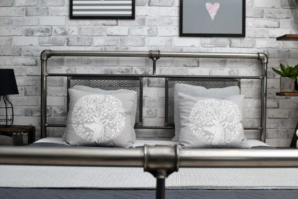 Szare metalowe łóżko industrialne w zbliżeniu na tle białej cegły i dekoracyjnych poduszek.