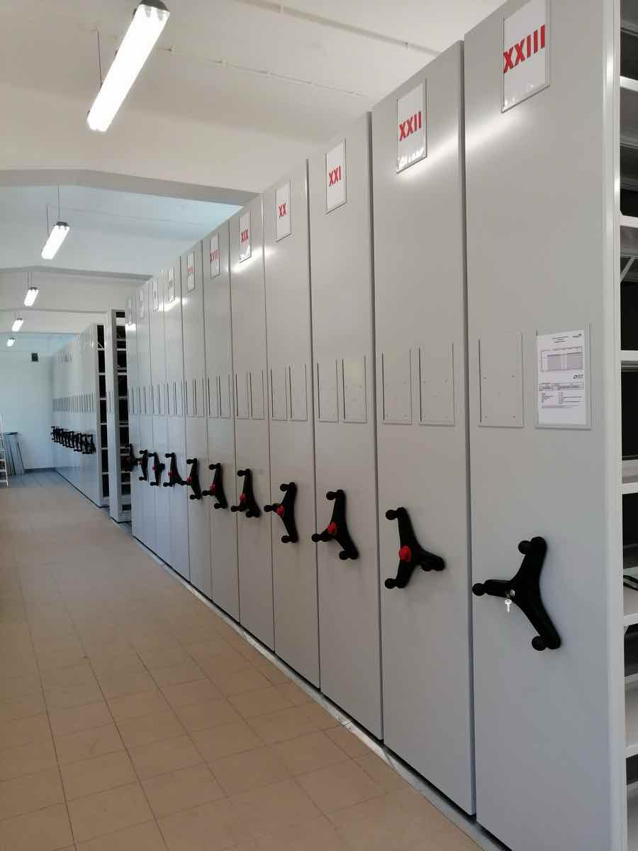 Regały przesuwne do archiwum z napędem mechanicznym, ustawione w długim ciągu w pomieszczeniu archiwalnym