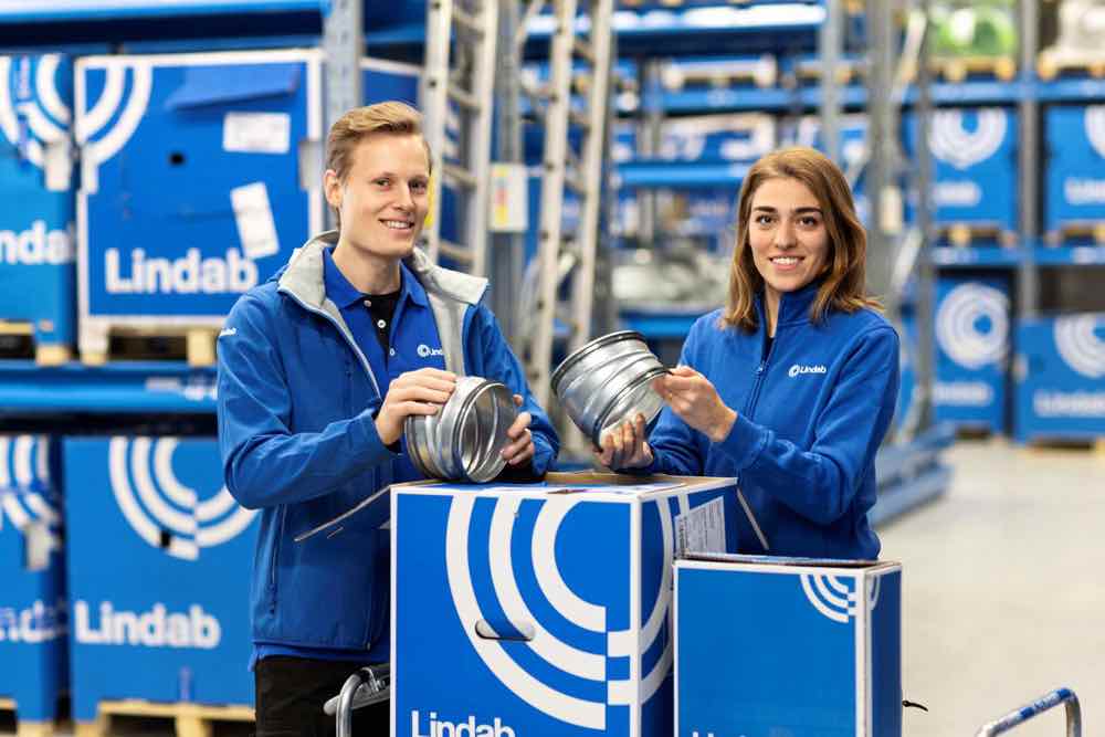 Eksperci Lindab w niebieskich strojach firmowych prezentują kształtki systemu wentylacji z fabryczną uszczelką