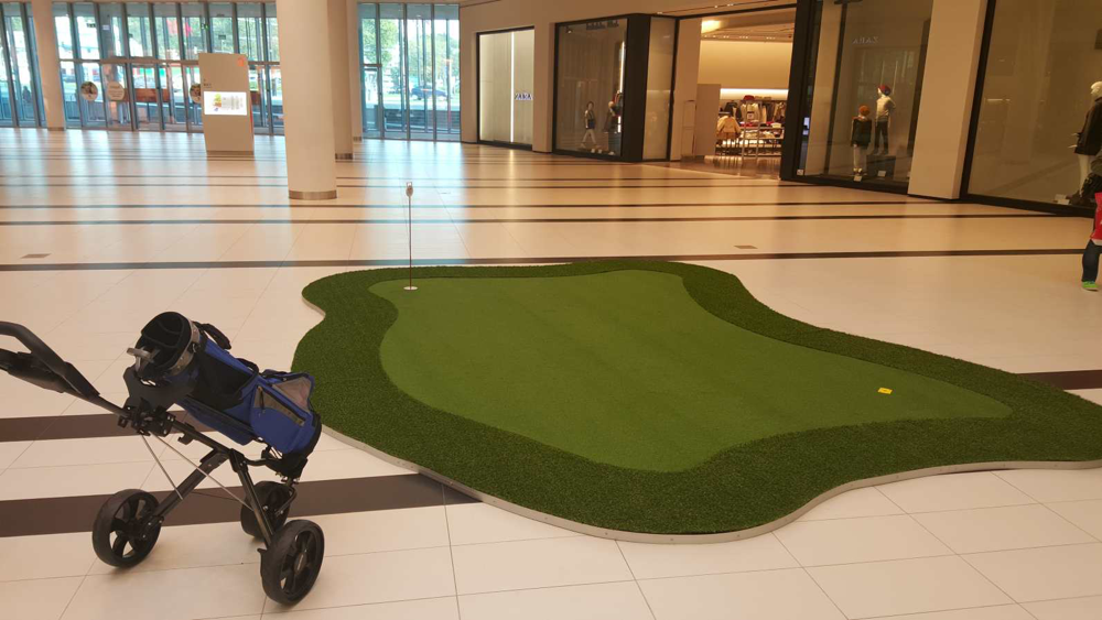 Green golfowy w galerii handlowej z polem do ćwiczenia uderzeń i wózkiem na kije golfowe.
